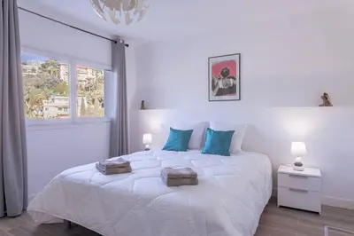 a white bedroom with a large white bed with blue pillows at Apartment With Panoramic Sea View, Walk To The Beach - Maréchal Juin by Halldis in Cannes