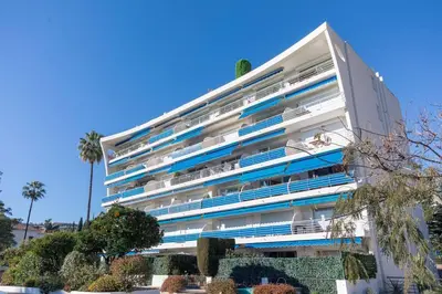 a large white building with blue windows and palm trees at Apartment With Panoramic Sea View, Walk To The Beach - Maréchal Juin by Halldis in Cannes